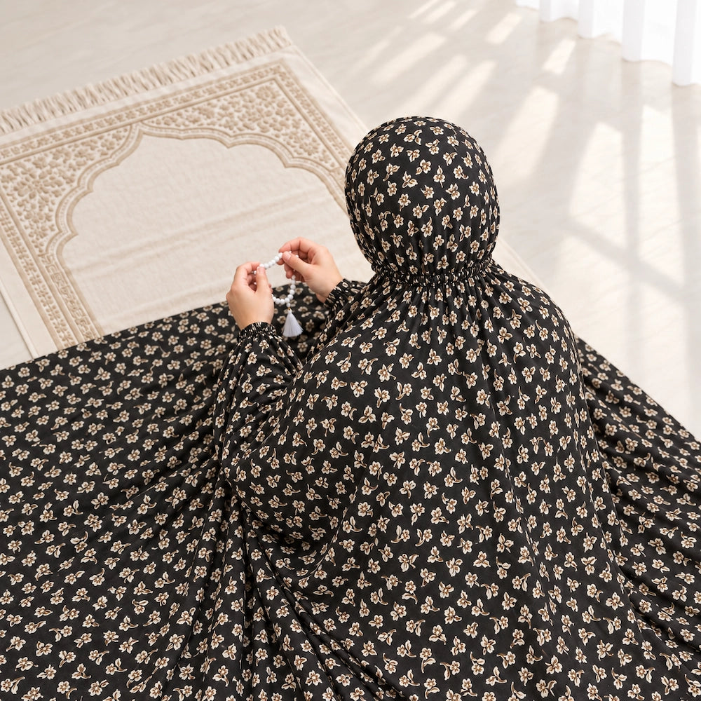 Woman holding prayer beads in a black namaz chaddar with tiny ivory floral sprigs, shown from behind on an ivory prayer mat.