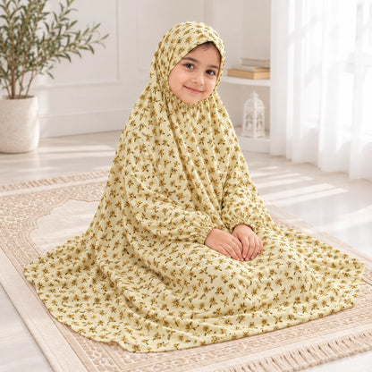 Girl seated on a prayer mat in a light yellow namaz chaddar with small mustard floral sprigs and gathered wrist cuffs.