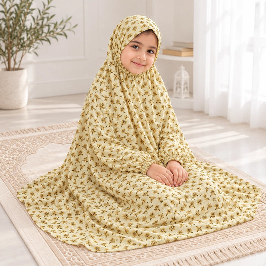 Girl seated on a prayer mat in a light yellow namaz chaddar with small mustard floral sprigs and gathered wrist cuffs.