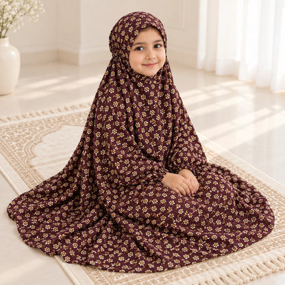 Girl seated on an ivory prayer mat wearing a maroon floral namaz chaddar with elasticated wrist cuffs.