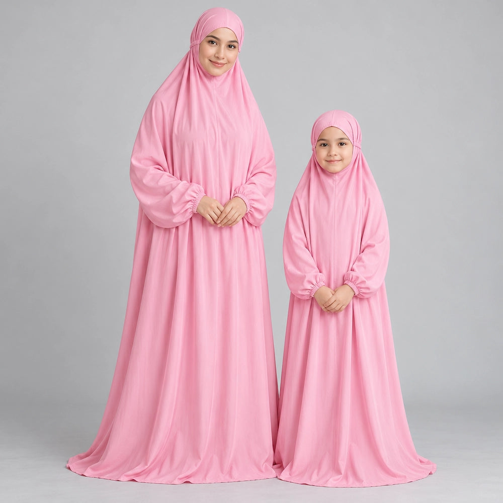 Mother and daughter standing in plain baby pink namaz chaddars with their hands gently clasped in front.