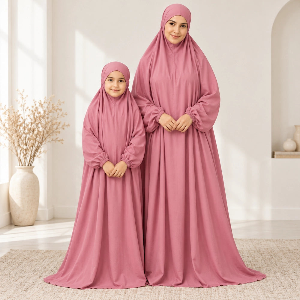 Mother and daughter standing with clasped hands in matching dusty rose namaz chaddars in a sunlit room.