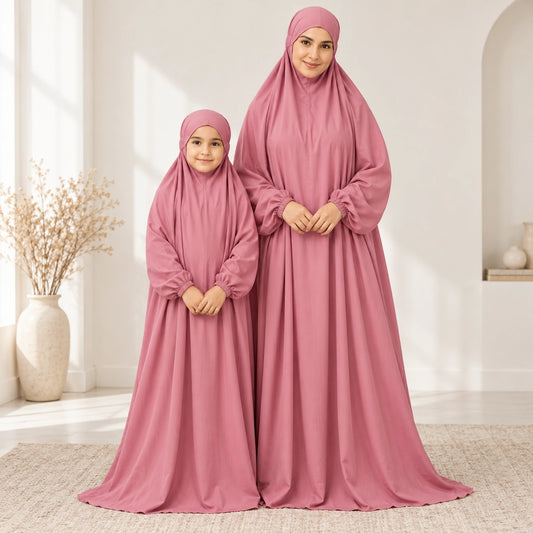 Mother and daughter standing with clasped hands in matching dusty rose namaz chaddars in a sunlit room.
