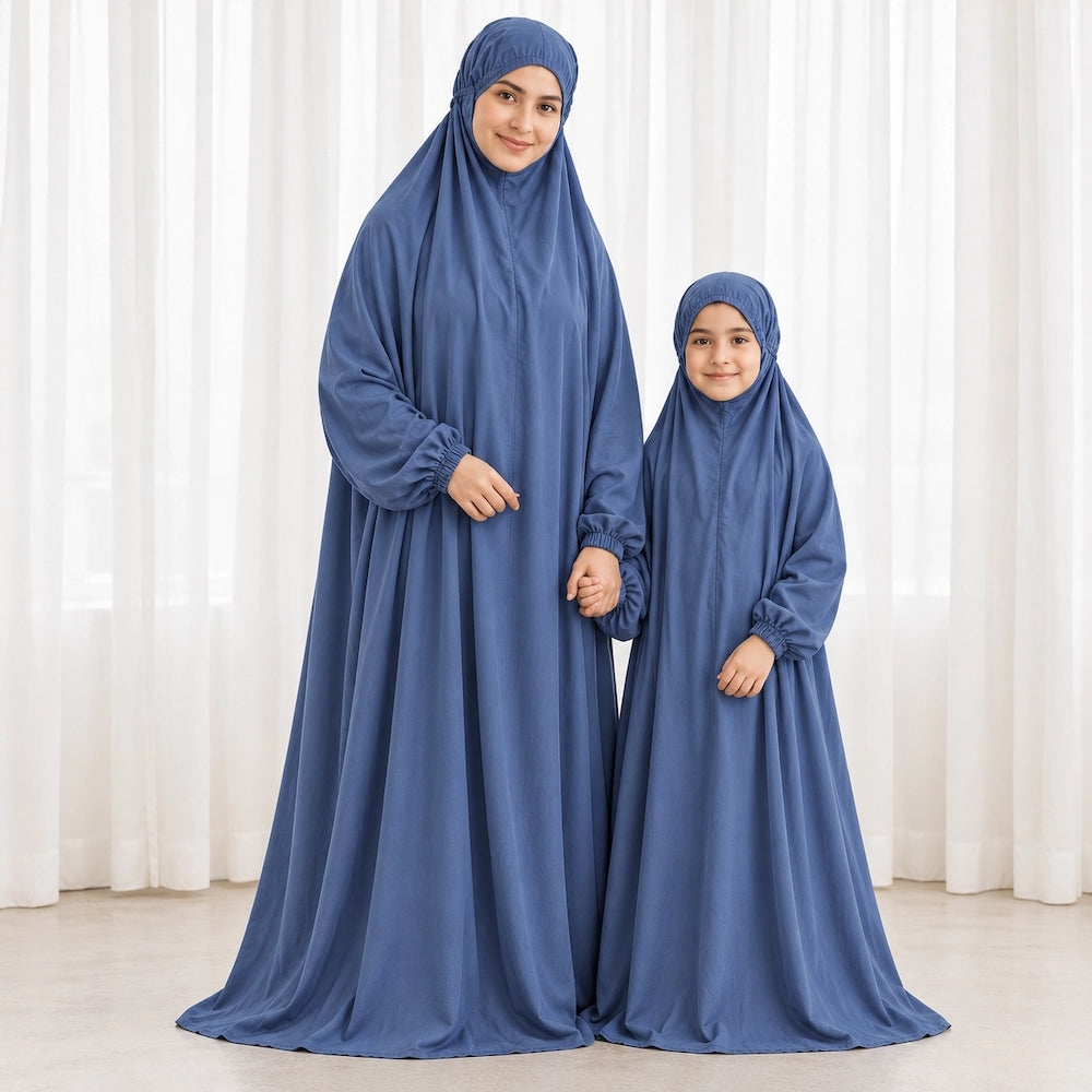 Mother and young daughter holding hands in matching dusty blue prayer chaddars beside white curtains.