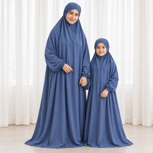 Mother and young daughter holding hands in matching dusty blue prayer chaddars beside white curtains.