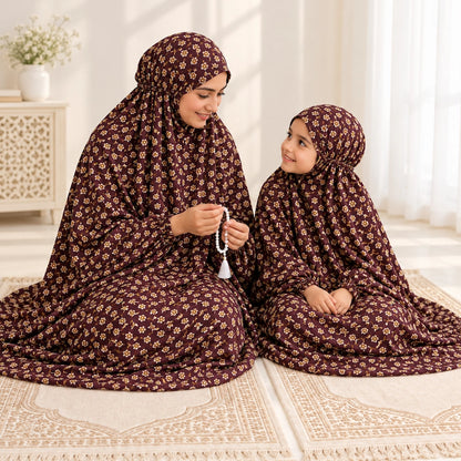 Mother and daughter seated on prayer mats in matching maroon floral namaz chaddars, with the mother holding prayer beads.