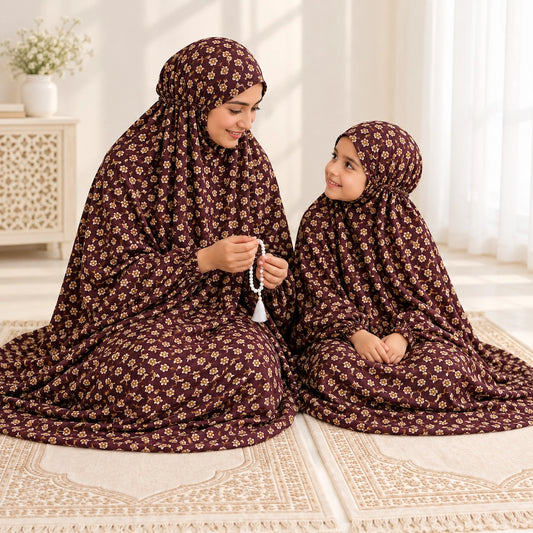 Mother and daughter seated on prayer mats in matching maroon floral namaz chaddars, with the mother holding prayer beads.