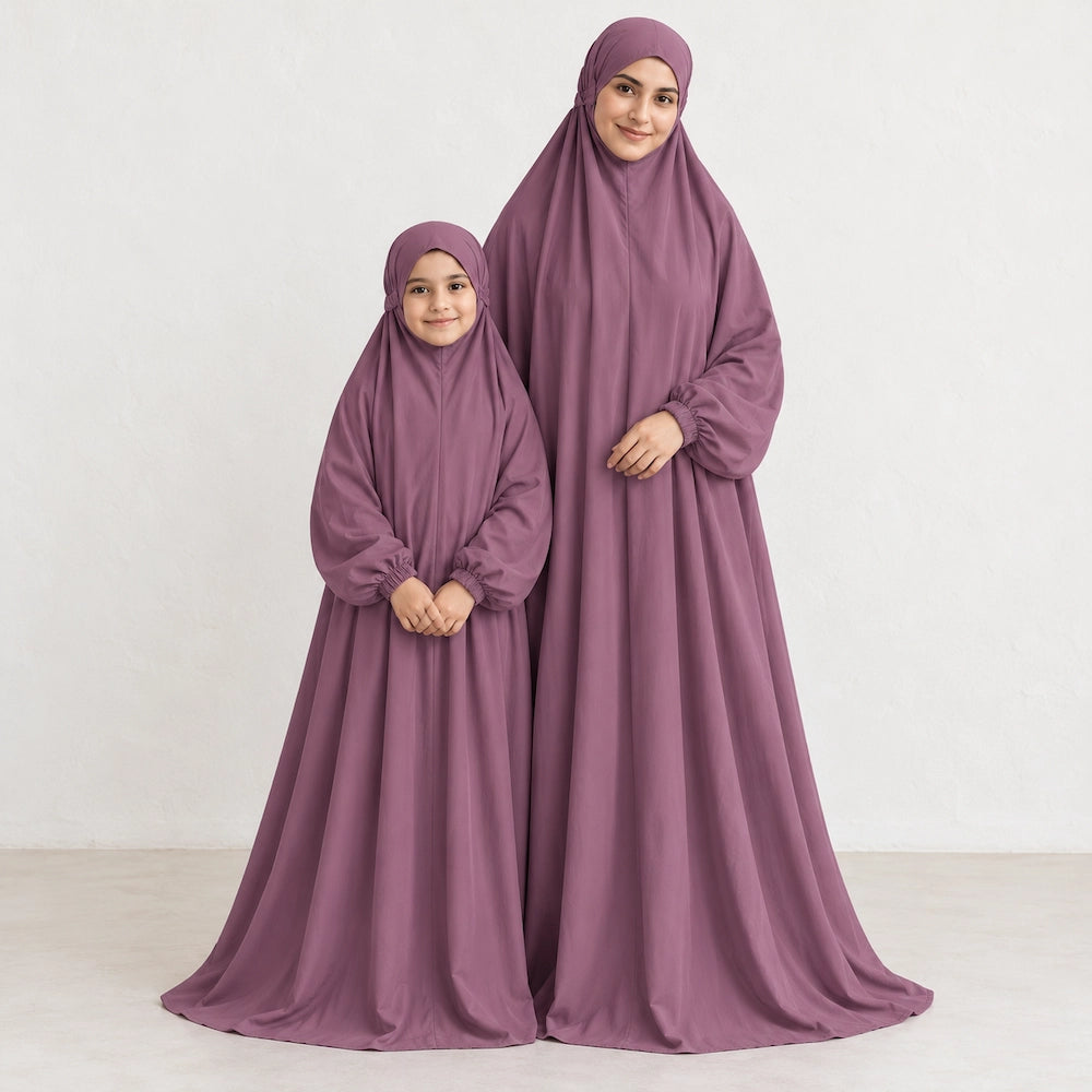 Daughter standing beside her mother in matching solid mauve namaz chaddars against a light textured wall.