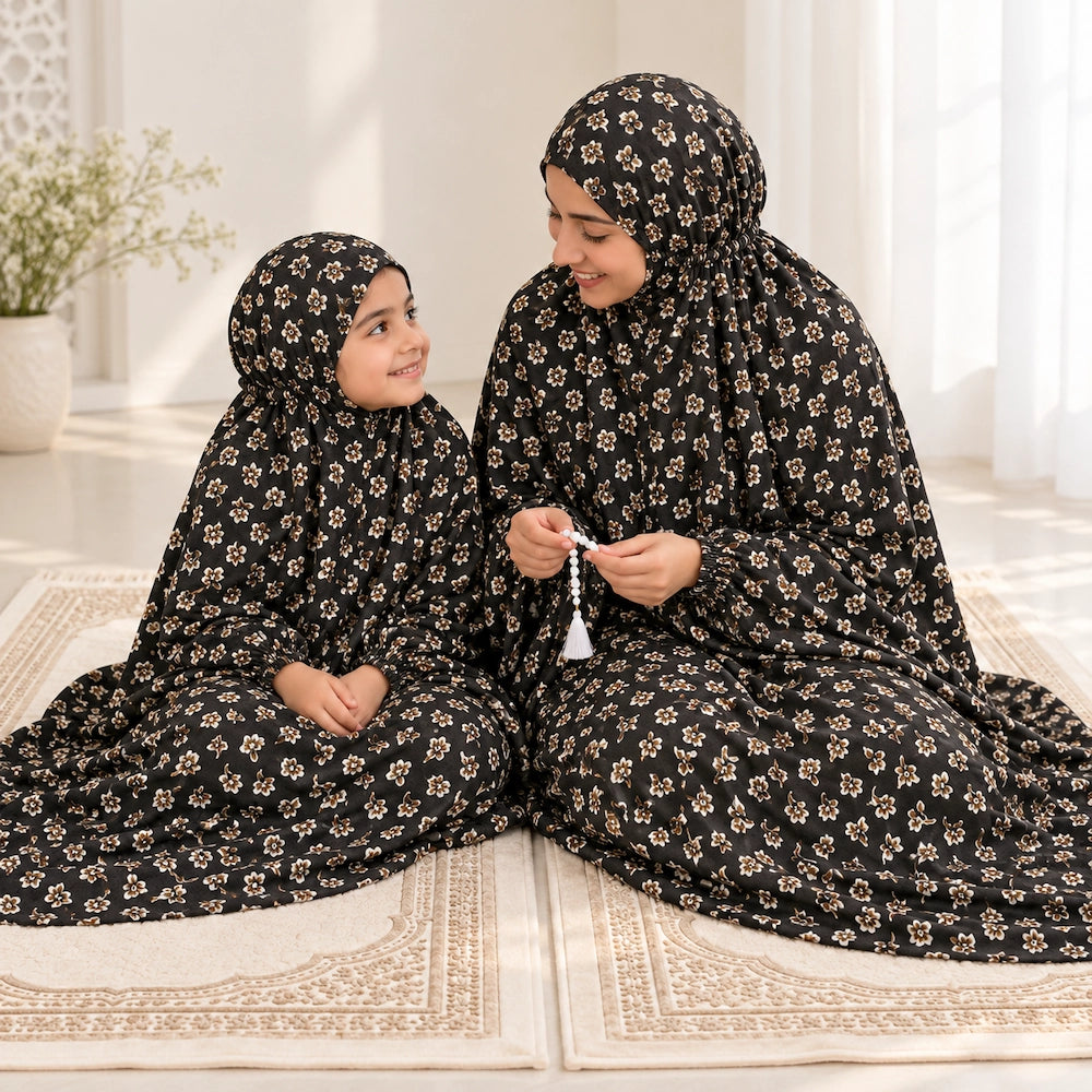 Mother and young daughter looking at each other while wearing matching black floral prayer chaddars on ivory prayer mats.