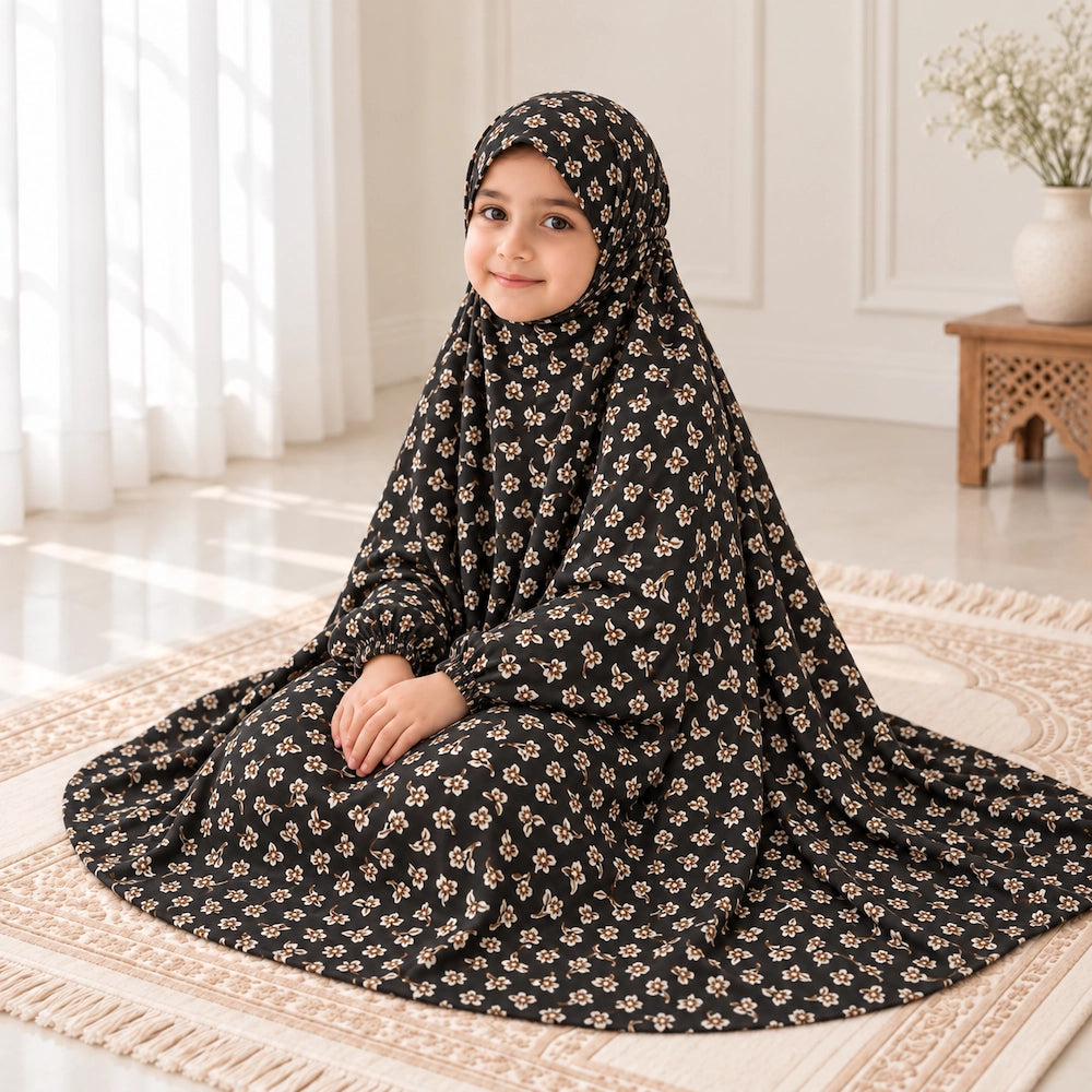 Young girl in a black namaz chaddar with small ivory floral sprigs, seated with her hands resting on her lap.