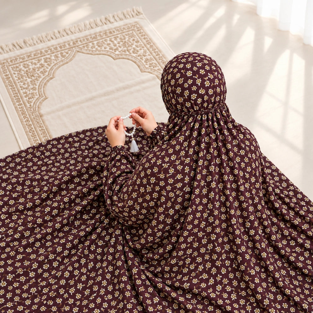 Woman seated on a prayer mat wearing a maroon small floral namaz chaddar with elasticated cuffs and head gathering.
