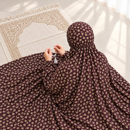 Woman seated on a prayer mat wearing a maroon small floral namaz chaddar with elasticated cuffs and head gathering.
