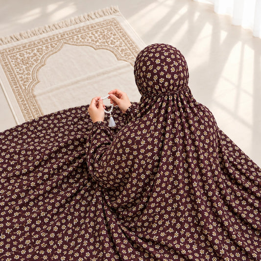 Woman seated on a prayer mat wearing a maroon small floral namaz chaddar with elasticated cuffs and head gathering.