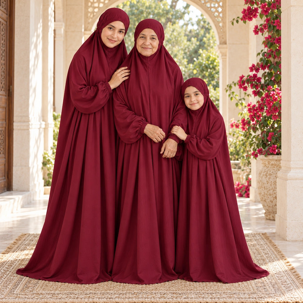 Mother and granddaughter standing close to grandmother in matching maroon namaz chaddars on a flower-lined veranda.