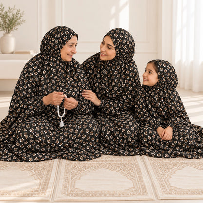 Three generations seated together on ivory prayer mats in matching black floral chaddars, with the grandmother holding prayer beads.