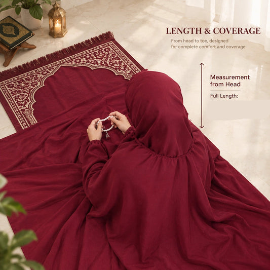 Woman wearing a plain maroon namaz chaddar, seated beside a prayer mat and holding prayer beads